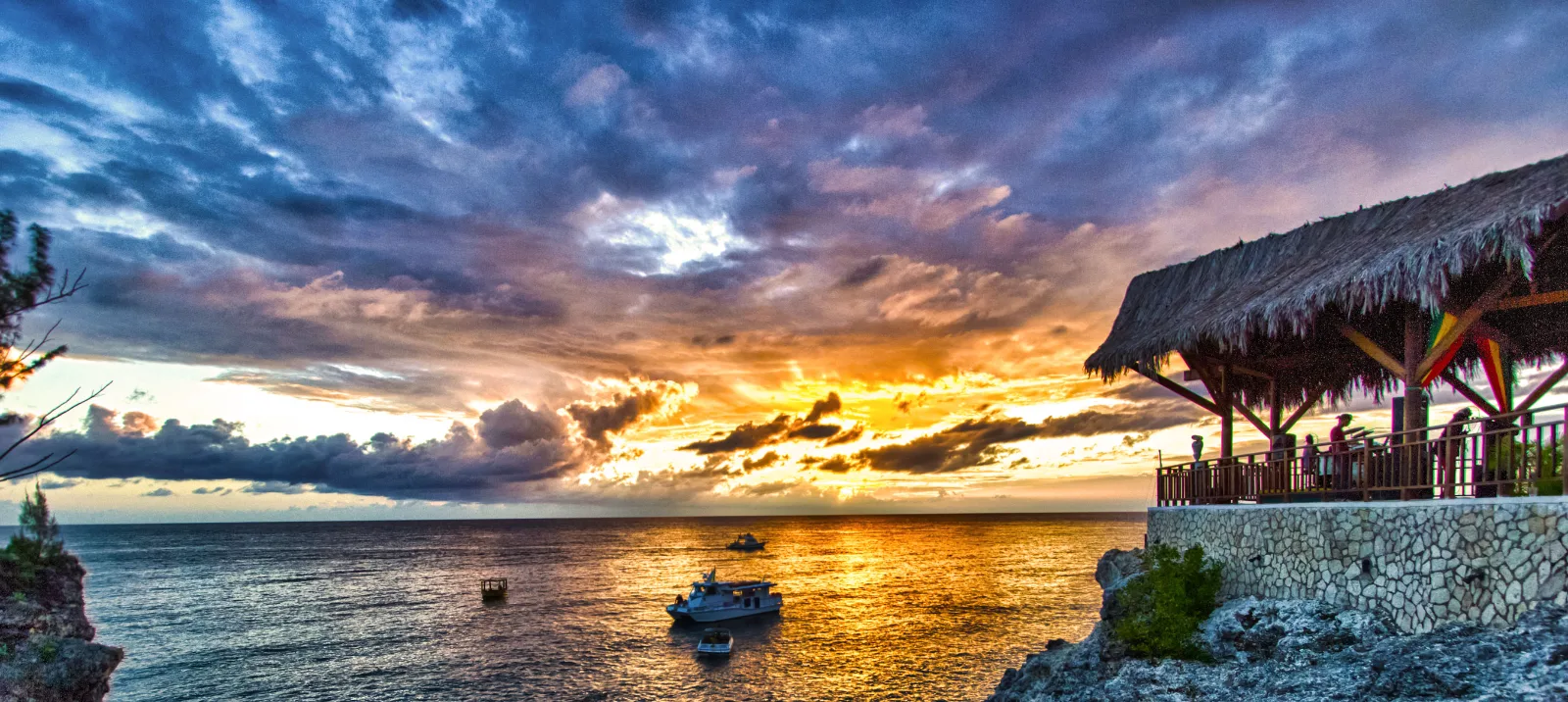 A thatched-roof cliffside pavilion overlooks boats on the glowing ocean beneath a dramatic sunset in Jamaica.