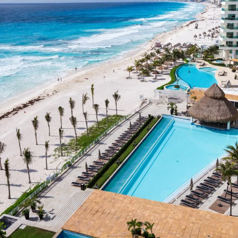 Resort swimming pools and sun loungers overlook a white-sand beach and turquoise ocean waters in Cancún, Mexico.