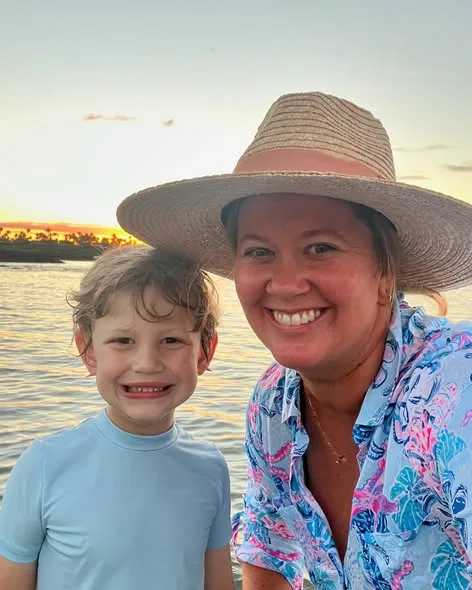 A smiling woman wearing a wide-brimmed straw hat poses with a young boy by the ocean at sunset.