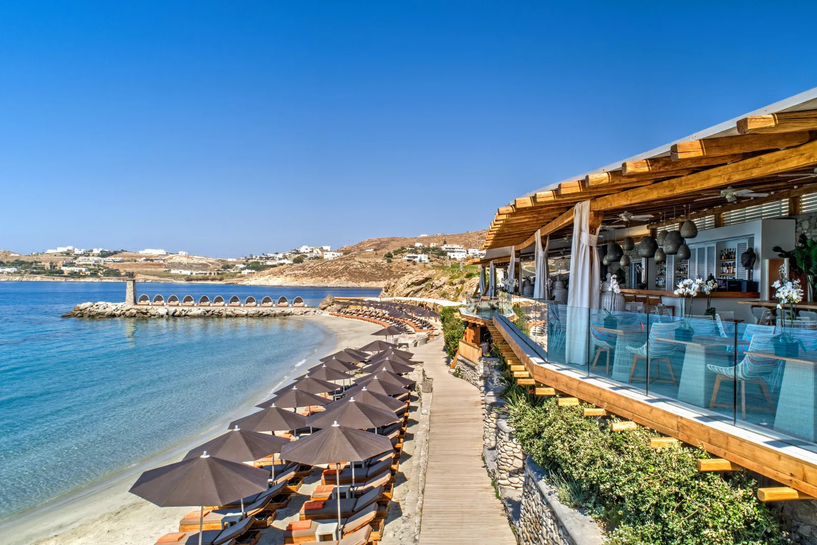 An open-air wooden restaurant terrace overlooks rows of umbrella-shaded loungers along a calm beach cove in Greece.