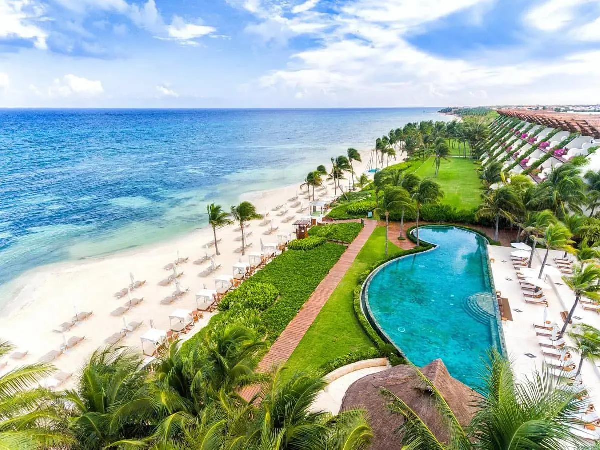 A curved swimming pool and palm-lined lawn overlook a white-sand beach beside turquoise ocean waters in Riviera Maya, Mexico.