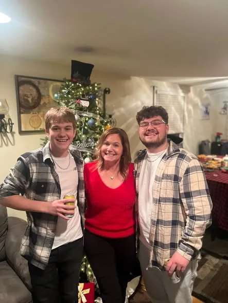 A smiling woman in a red sweater poses with two young men in plaid shirts before an illuminated Christmas tree inside a home.