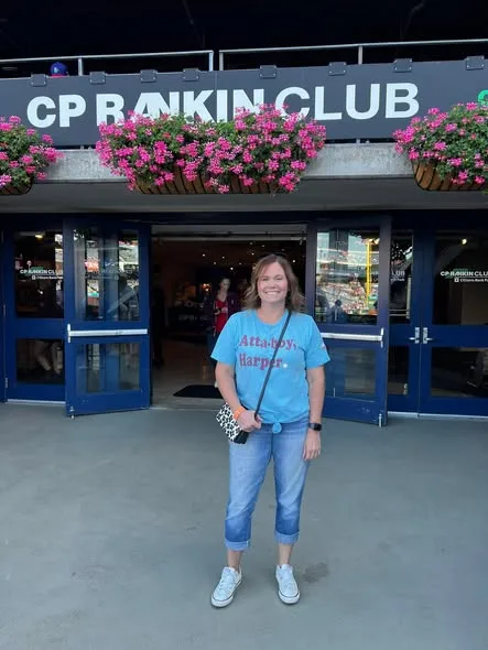 A smiling woman in a blue t-shirt stands outside a stadium club entrance adorned with hanging pink flower boxes.