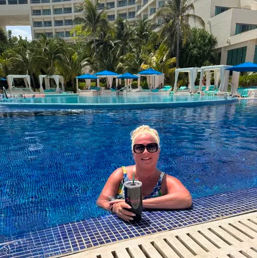 A woman in sunglasses relaxes in a resort swimming pool with a drink, backed by poolside cabanas and palm trees.