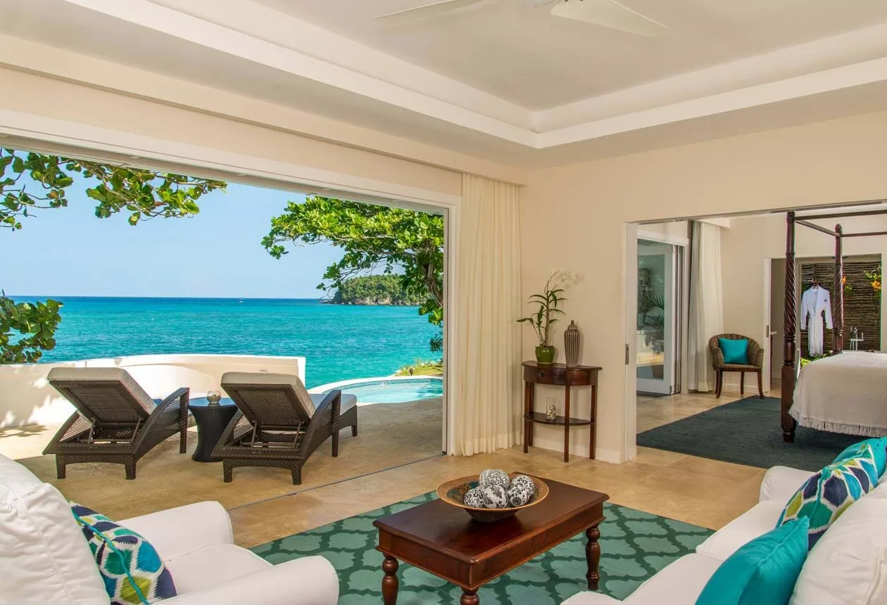 A bright cottage living room opens onto a sun terrace with loungers overlooking the turquoise sea in Jamaica.