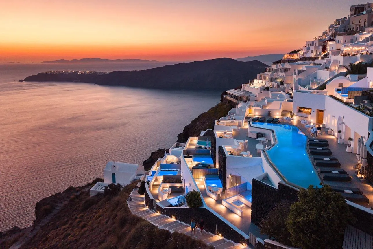 Tiered whitewashed villas with illuminated swimming pools sit along the cliffs of Santorini, overlooking the sea at sunset.