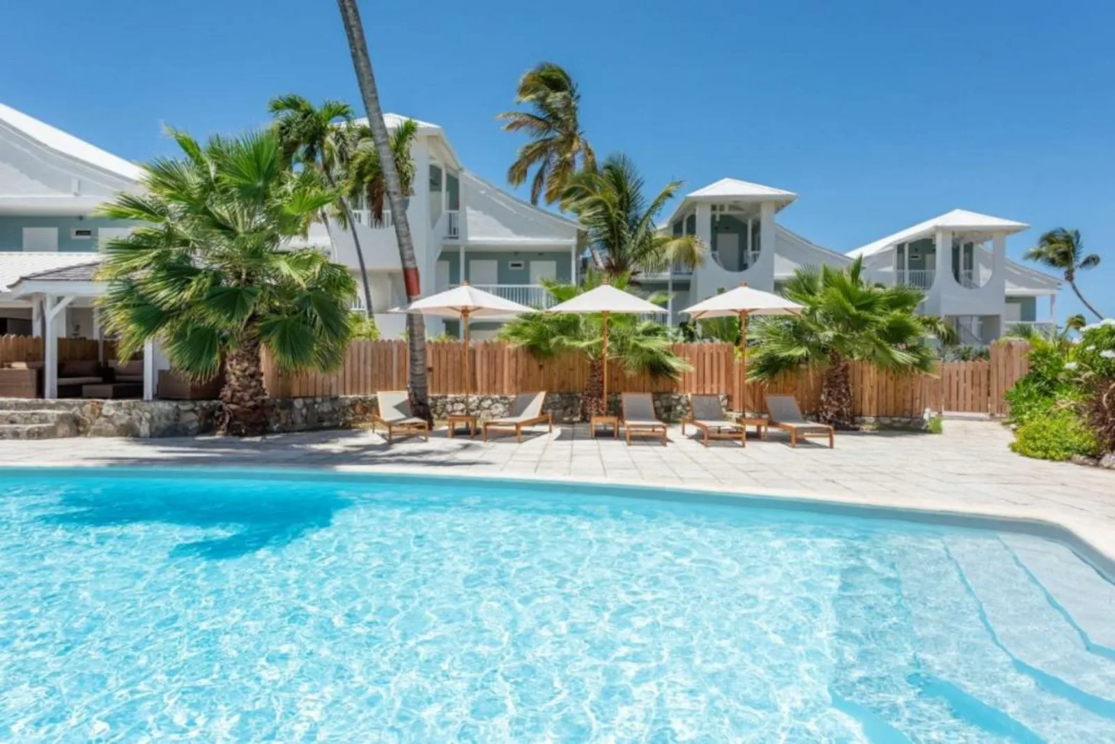 Sun loungers with parasols stand beside a turquoise swimming pool, surrounded by palm trees and white villas in St. Martin.
