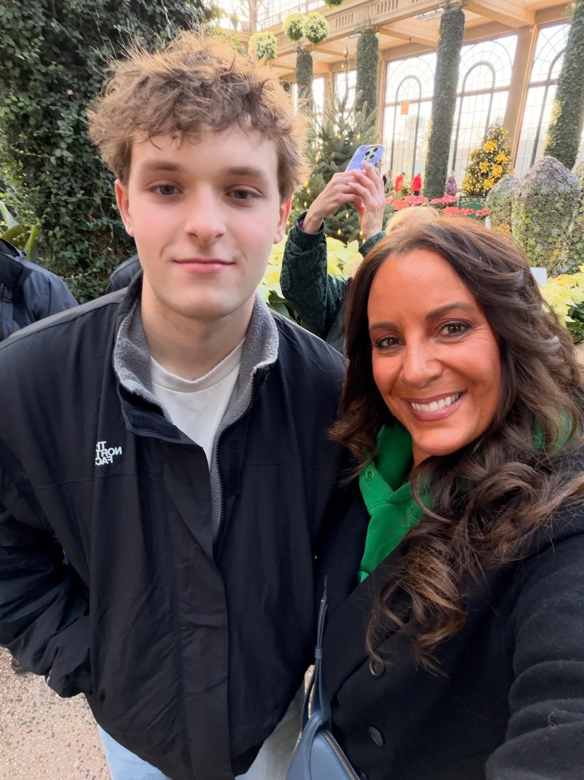A smiling woman and a young man pose for a selfie inside a sunlit botanical conservatory filled with lush plants.