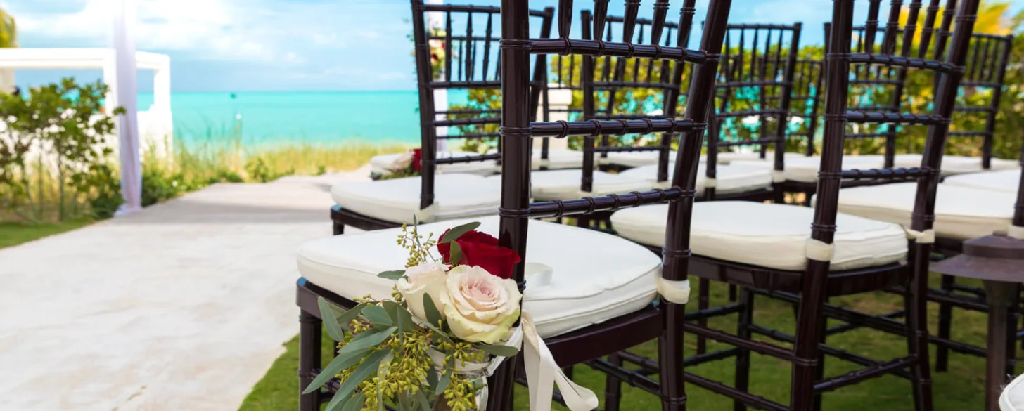 Dark wooden Chiavari chairs with white cushions and floral arrangements line an aisle overlooking a turquoise tropical ocean.