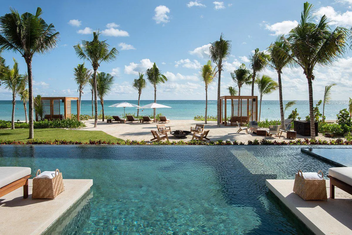 An infinity pool terrace overlooks a palm-studded beach with wooden cabanas and the turquoise ocean in Riviera Maya, Mexico.