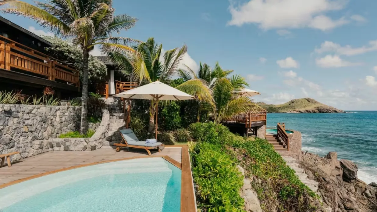 A private plunge pool and sun lounger overlook a rocky coastline and turquoise ocean from a tropical villa terrace.
