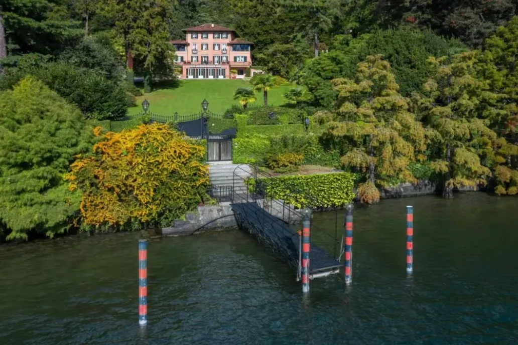 A private boat dock with striped mooring poles leads up to a pink waterfront villa surrounded by lush terraced gardens.
