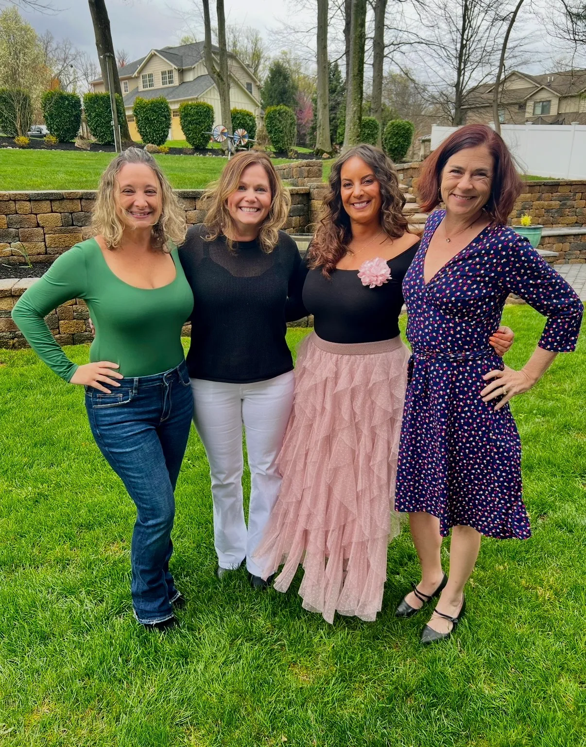 Four smiling women pose together on a green lawn in a suburban backyard bordered by stone walls and trees.