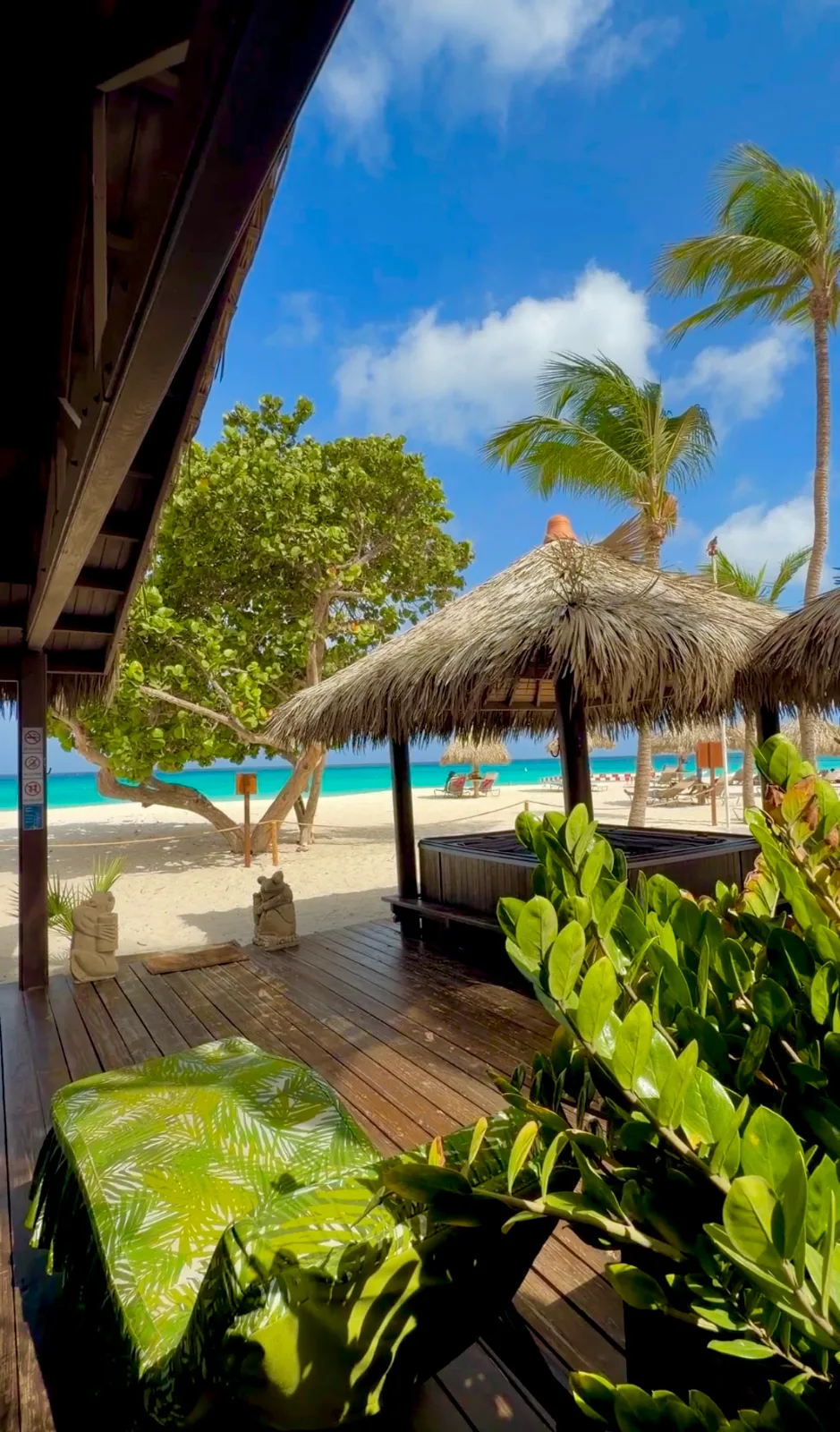 A shaded wooden deck with a massage bed and thatched palapa overlooks a white-sand beach and turquoise sea in Aruba.