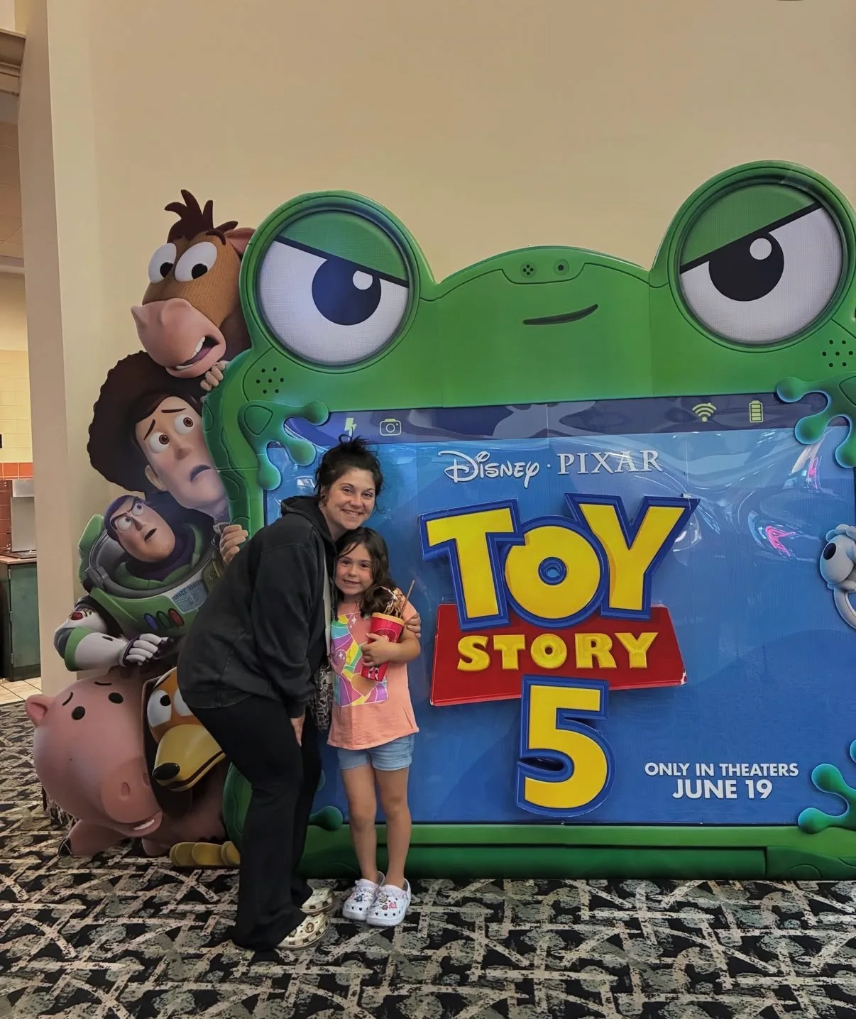 A woman and young girl pose beside a large Toy Story 5 standee in a carpeted cinema lobby.