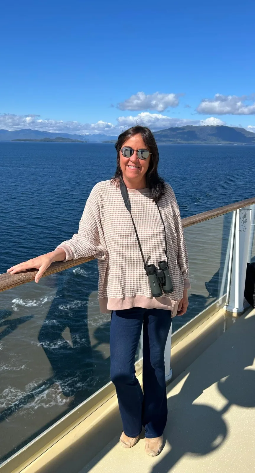 A smiling woman with binoculars stands on a cruise ship deck overlooking deep blue ocean waters and distant coastal mountains.