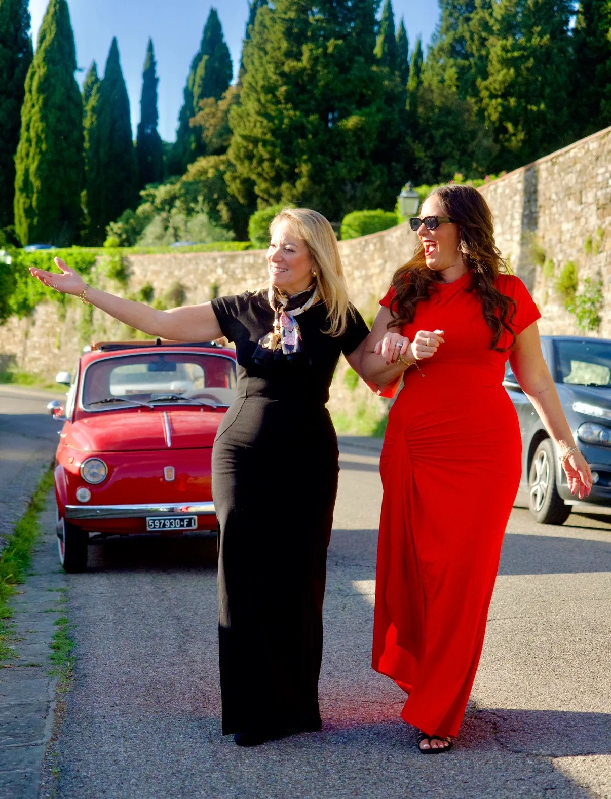 Two women in long dresses walk arm in arm past a vintage red Fiat on a road lined with cypress trees.