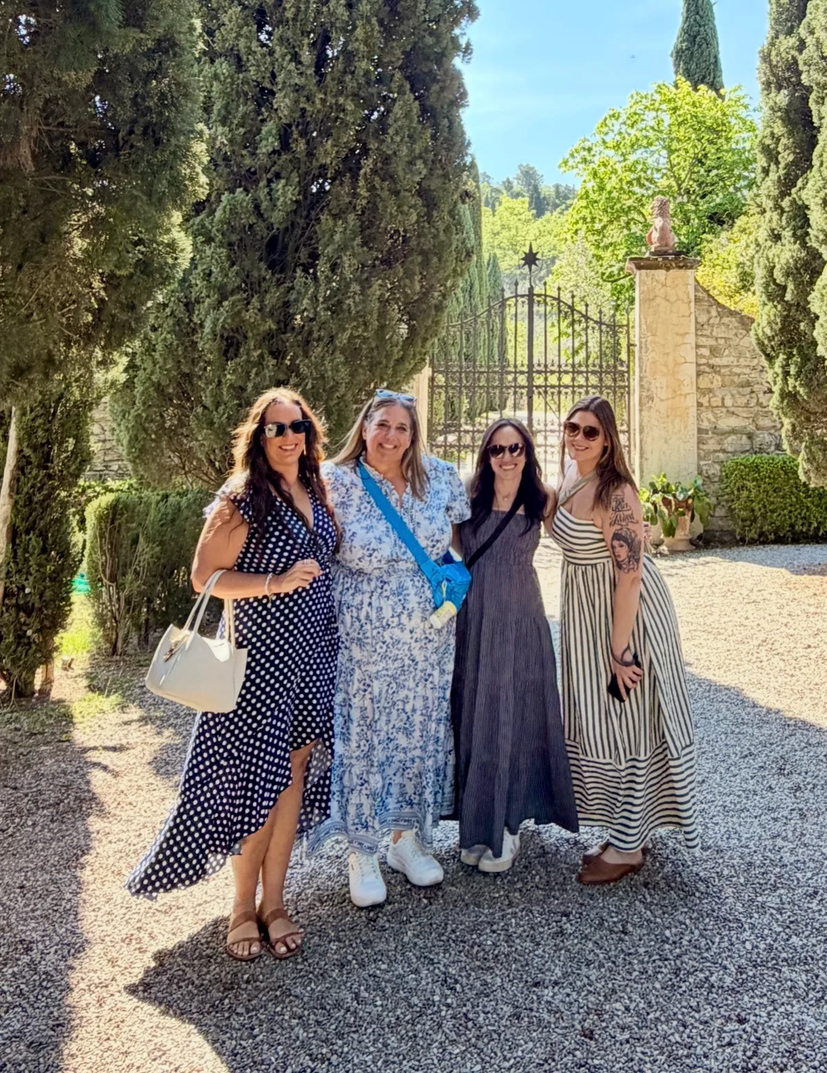 Four women in summer dresses smile together on a gravel driveway outside an estate gate surrounded by tall cypress trees.