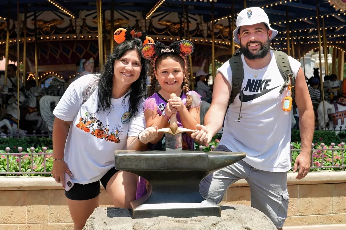 A smiling family poses around the Sword in the Stone in front of an ornate carousel at a Disney theme park.