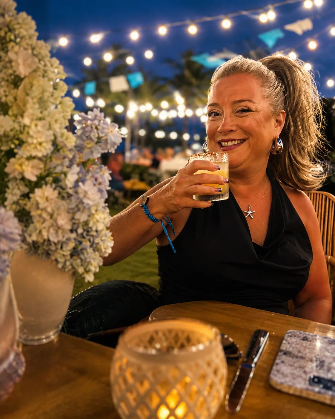 A smiling woman holds a cocktail at an outdoor dinner table illuminated by string lights and candlelight.