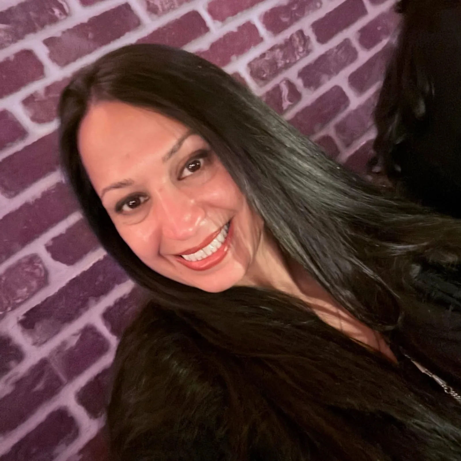 A smiling woman with long dark hair poses indoors in front of a purple brick-patterned wall.