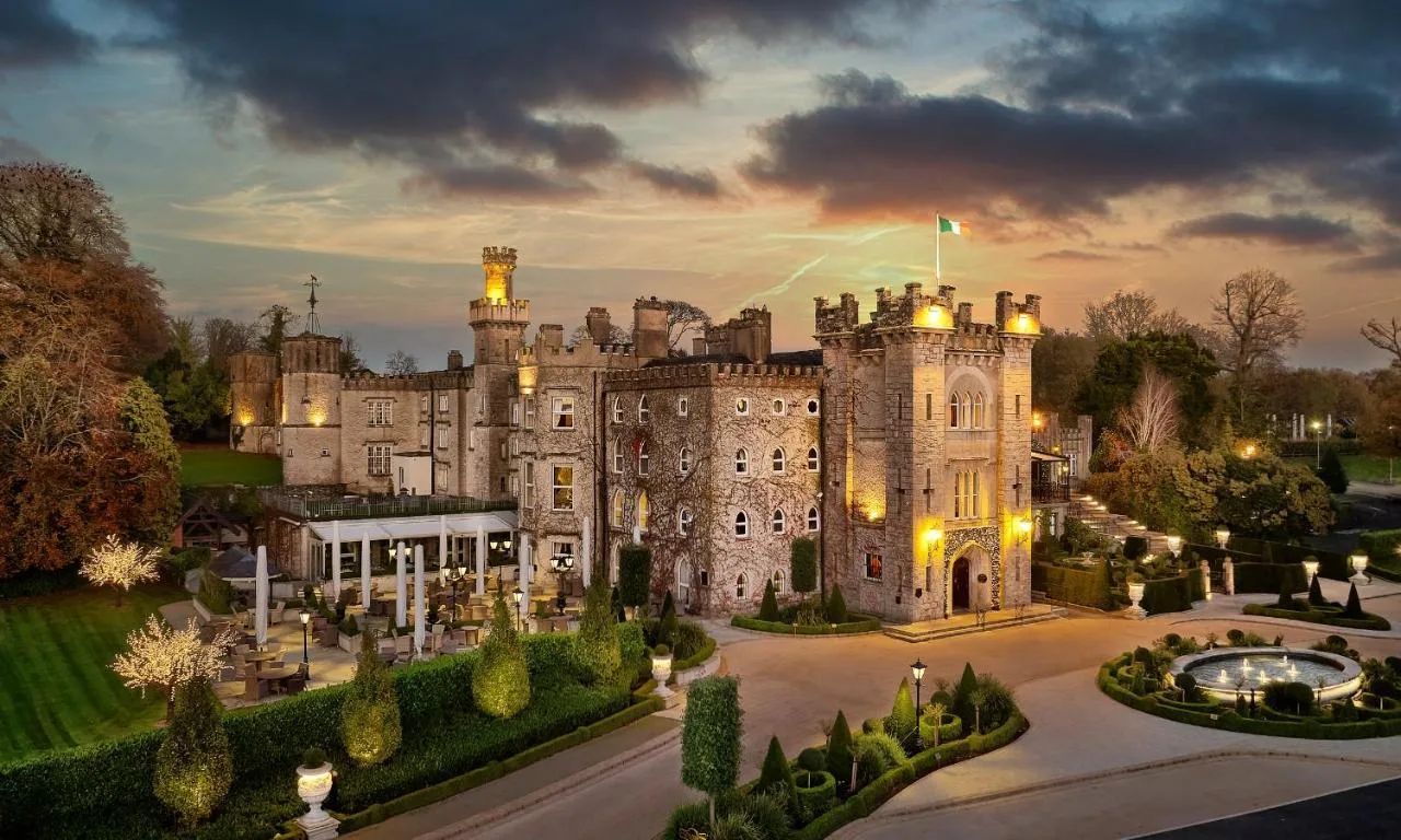 An illuminated historic stone castle hotel stands surrounded by manicured gardens and a fountain courtyard beneath a dramatic twilight sky in Ireland.