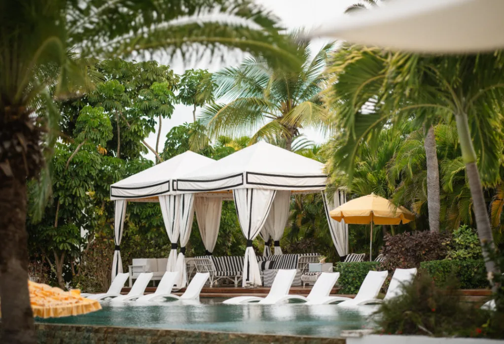 White canopied cabanas and poolside loungers sit beside a swimming pool surrounded by lush palm trees at an Anguilla resort.