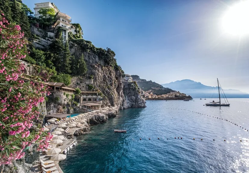 Sun loungers and parasols line a rocky seaside terrace below steep cliffs along the Amalfi Coast, Italy.