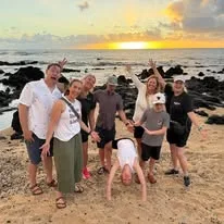 A smiling family poses together on a sandy beach with dark coastal rocks as the sun sets over the ocean.