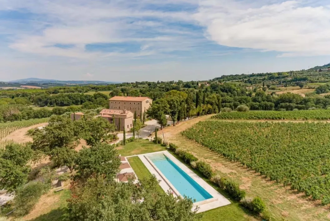 A stone villa estate and a long swimming pool sit surrounded by lush vineyards and rolling green countryside.