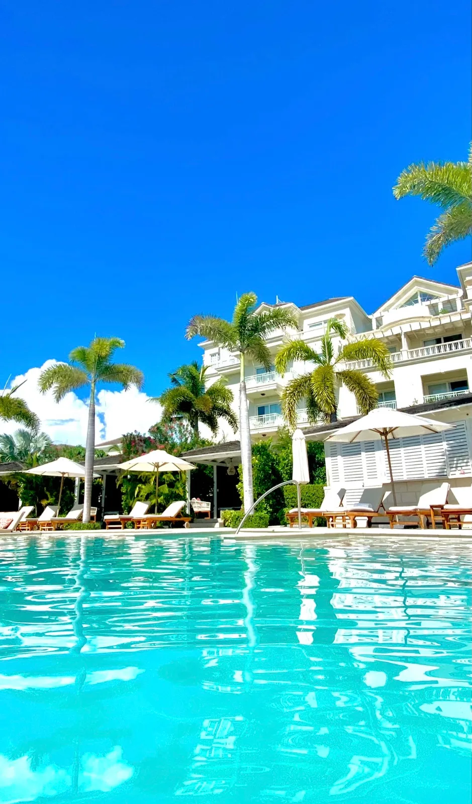 A turquoise swimming pool sits surrounded by lounge chairs, sun umbrellas, and palm trees before a white resort in Turks & Caicos.