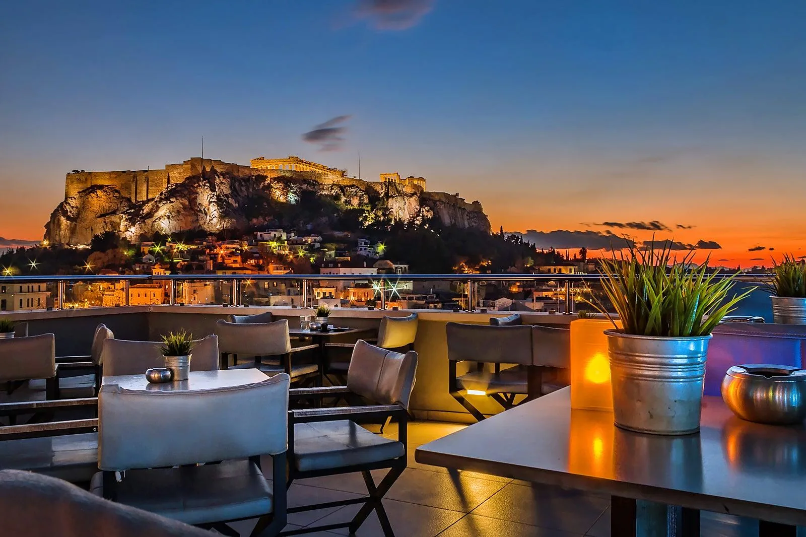 A rooftop terrace with dining tables overlooks the illuminated Acropolis in Athens against a colorful sunset sky.