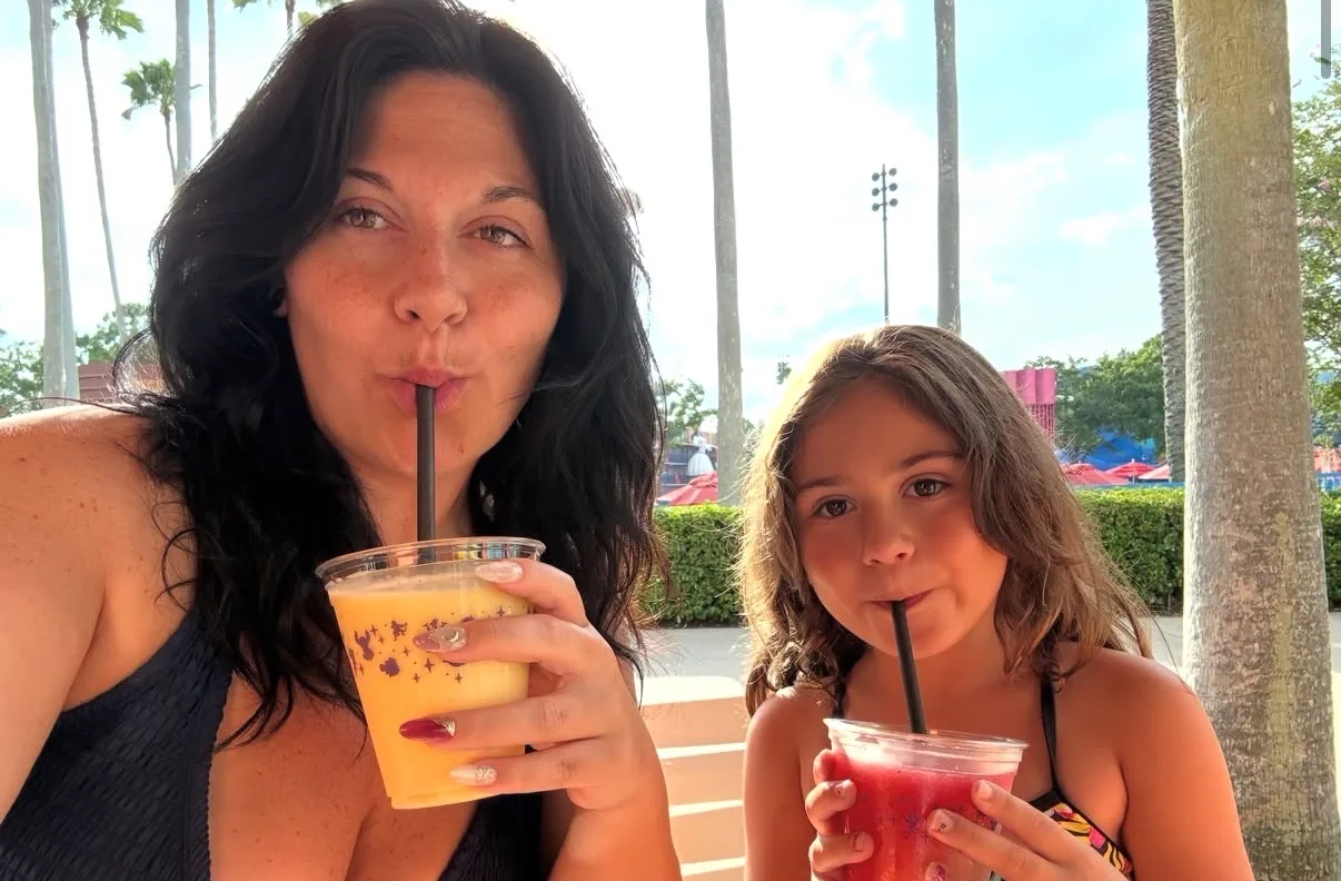 A woman and a young girl drink colorful iced fruit smoothies through straws while sitting outdoors beside tall palm trees.