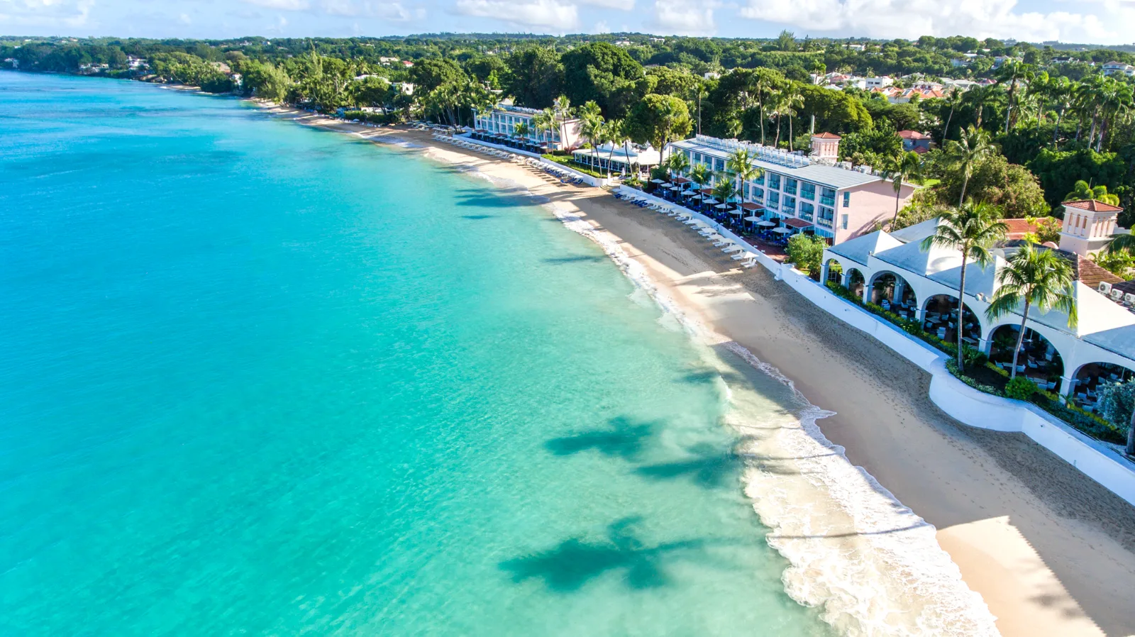 Beachfront resort buildings line a sandy shore beside clear turquoise ocean waters along the coast of Barbados.