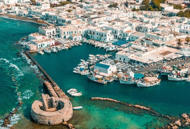 Fishing boats moored in a turquoise harbor beside a stone fortress ruin and whitewashed coastal village in Greece.