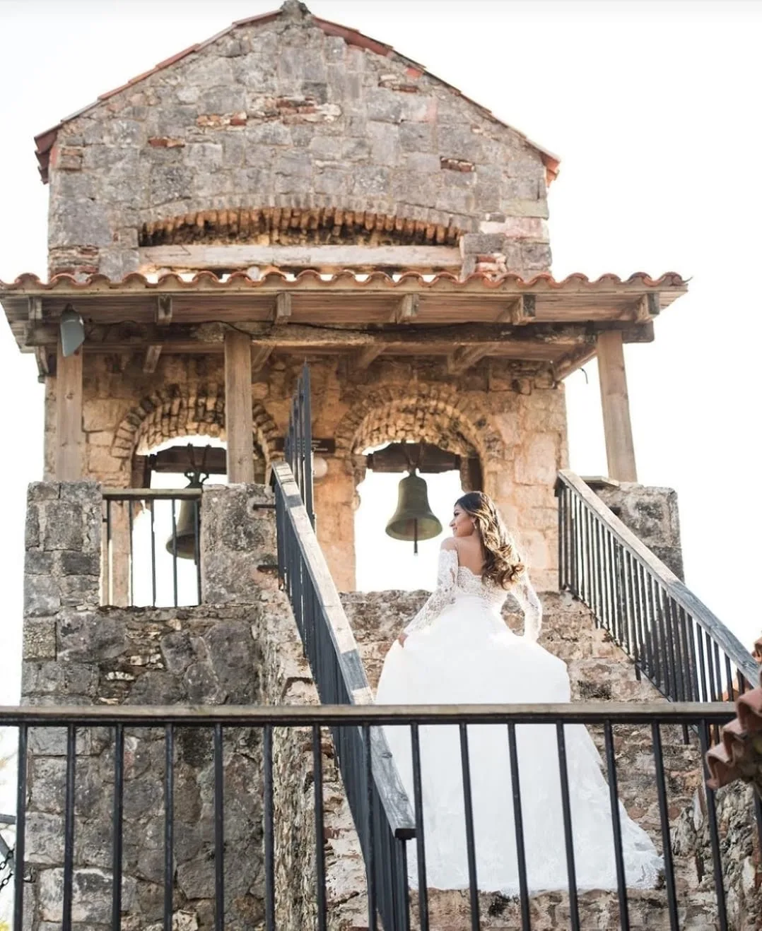 A bride in a white lace gown ascends stone steps toward a rustic bell tower with arched belfries.