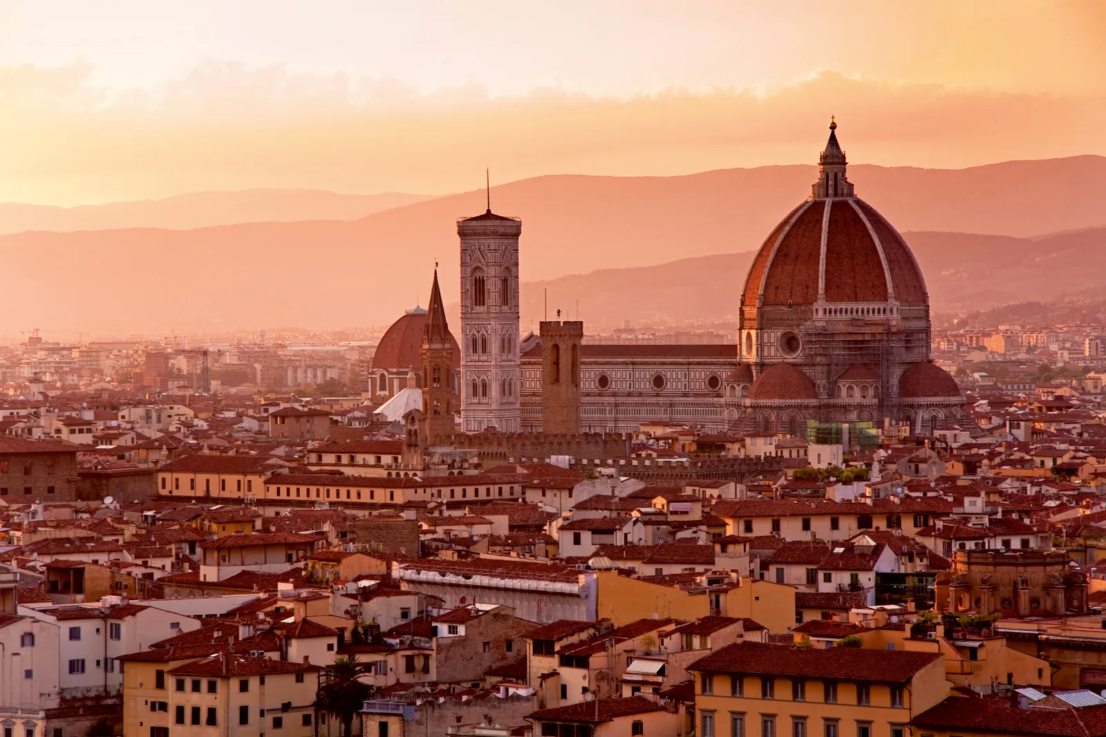 Florence Cathedral and Giotto's Campanile rise above terracotta rooftops and distant hills under a warm sunset glow in Florence, Italy.