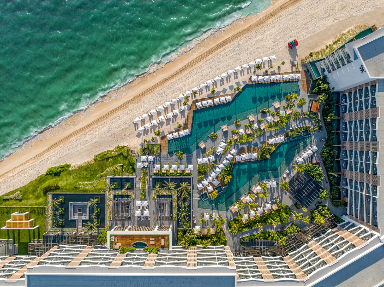 Swimming pools and white umbrellas at a beachfront resort border a sandy coastline and turquoise ocean in Cancun, Mexico.