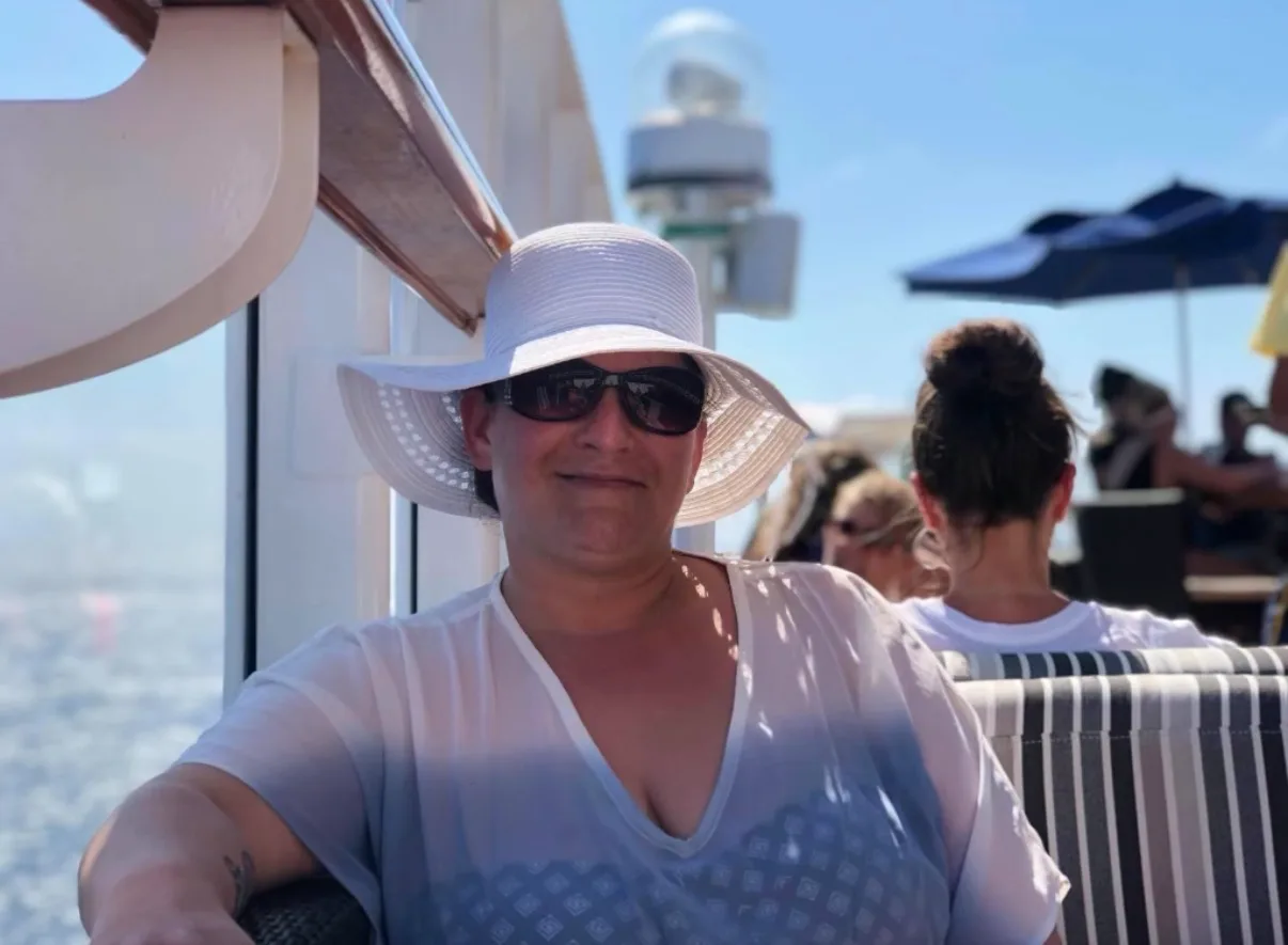 A woman wearing a white sunhat and sunglasses relaxes on the sunny open deck of a cruise ship.