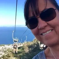 A smiling woman wearing sunglasses rides an open-air chairlift overlooking a coastal hillside and deep blue sea.