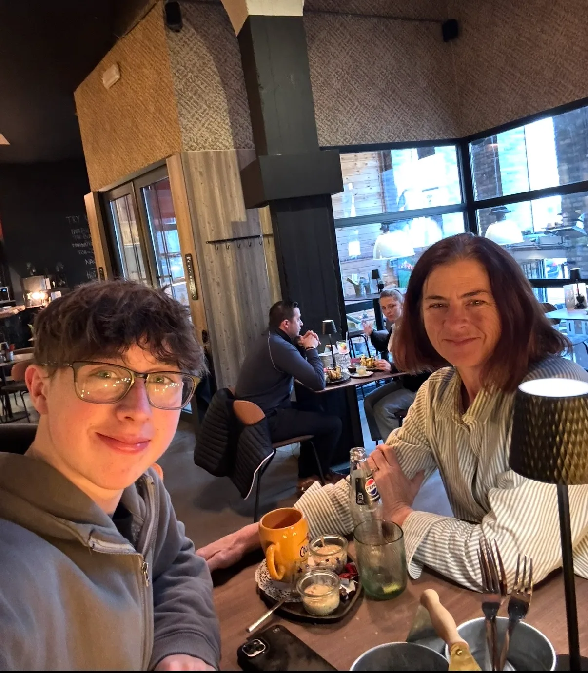 A young man wearing glasses and a woman smile at their table inside a cozy restaurant with drinks and desserts.