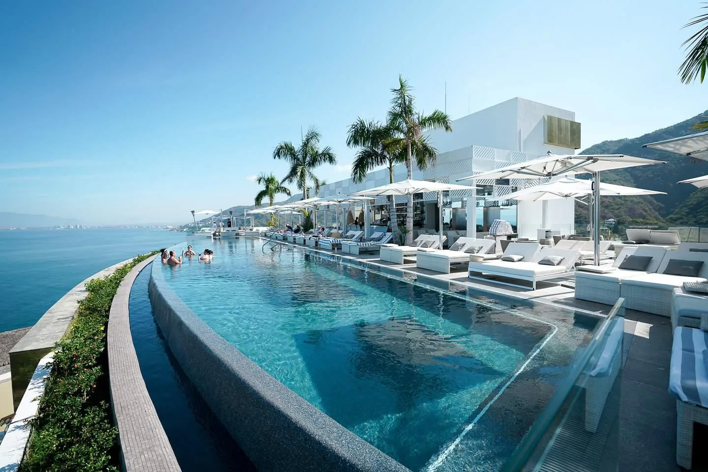 A curved rooftop infinity pool lined with white sun loungers overlooks the ocean and mountains in Puerto Vallarta, Mexico.