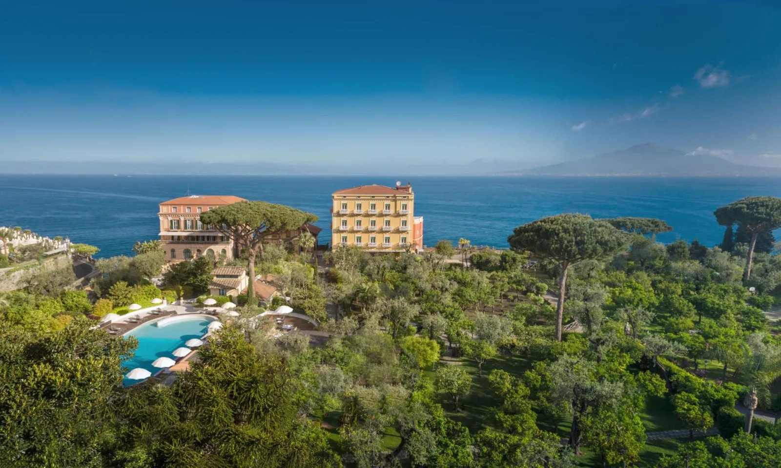 Historic coastal hotel buildings and a swimming pool nestled in lush gardens overlook the sea and Mount Vesuvius in Italy.