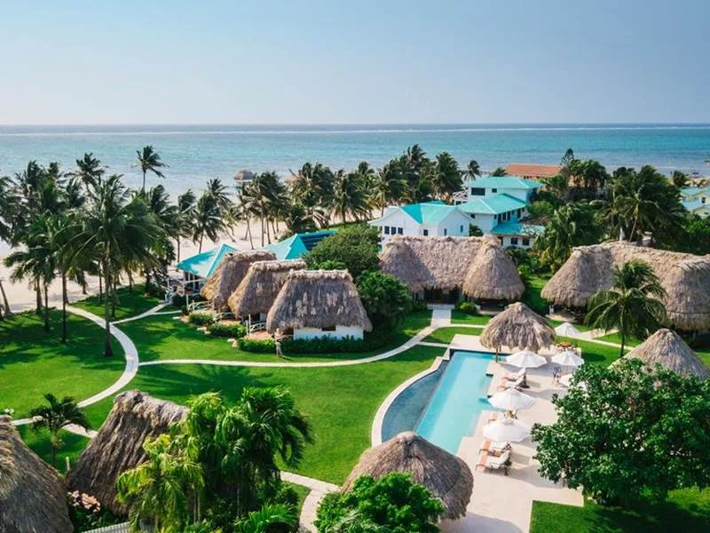 Thatched-roof resort bungalows and a swimming pool border a white sand beach and turquoise ocean in Belize.