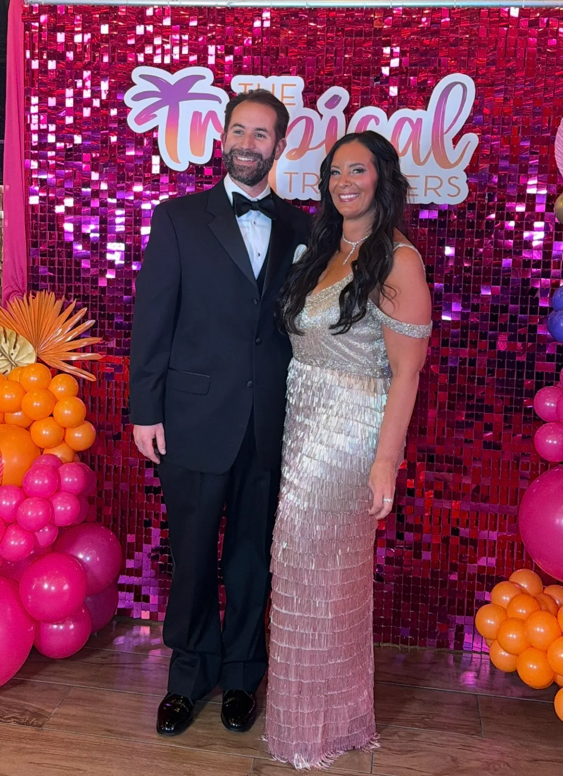 A formally dressed couple poses together in front of a sparkling magenta sequin backdrop decorated with colorful balloons.