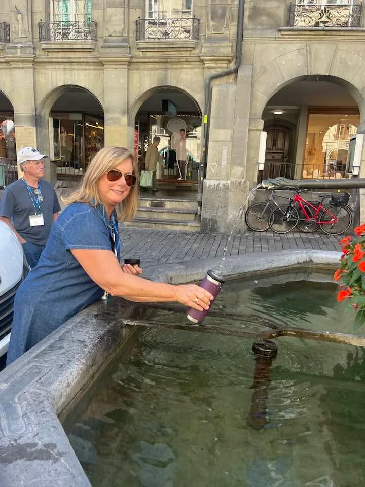 A woman fills a water bottle at a public stone fountain along a cobblestone street with historic arcades.