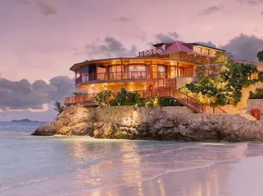 An illuminated multi-level villa sits on a rocky cliff above a sandy beach and calm ocean at dusk in St. Barts.