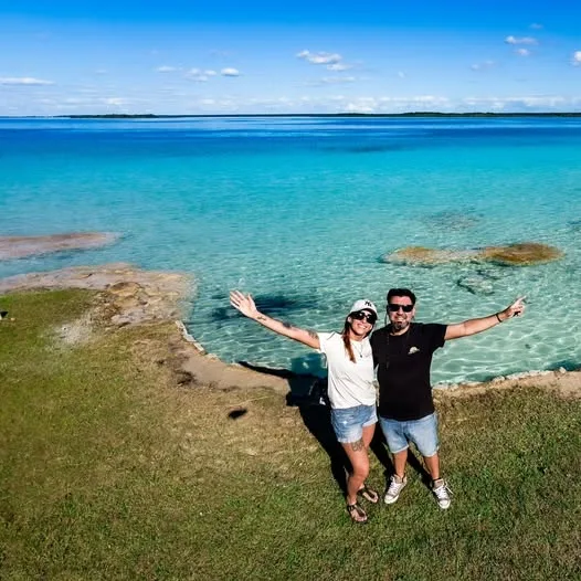 A couple stands with arms outstretched on a grassy shoreline overlooking clear turquoise waters under a sunny blue sky.