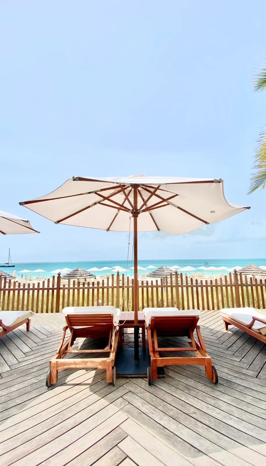 Wooden lounge chairs sit beneath a white sun umbrella on a deck overlooking a turquoise beach in Turks and Caicos.