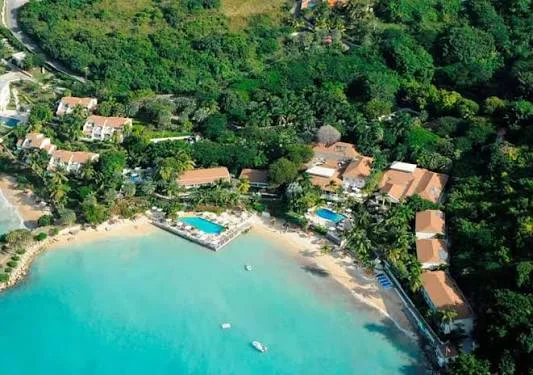 A beachfront resort with swimming pools and terracotta roofs sits along a turquoise bay in Antigua, surrounded by lush tropical forest.